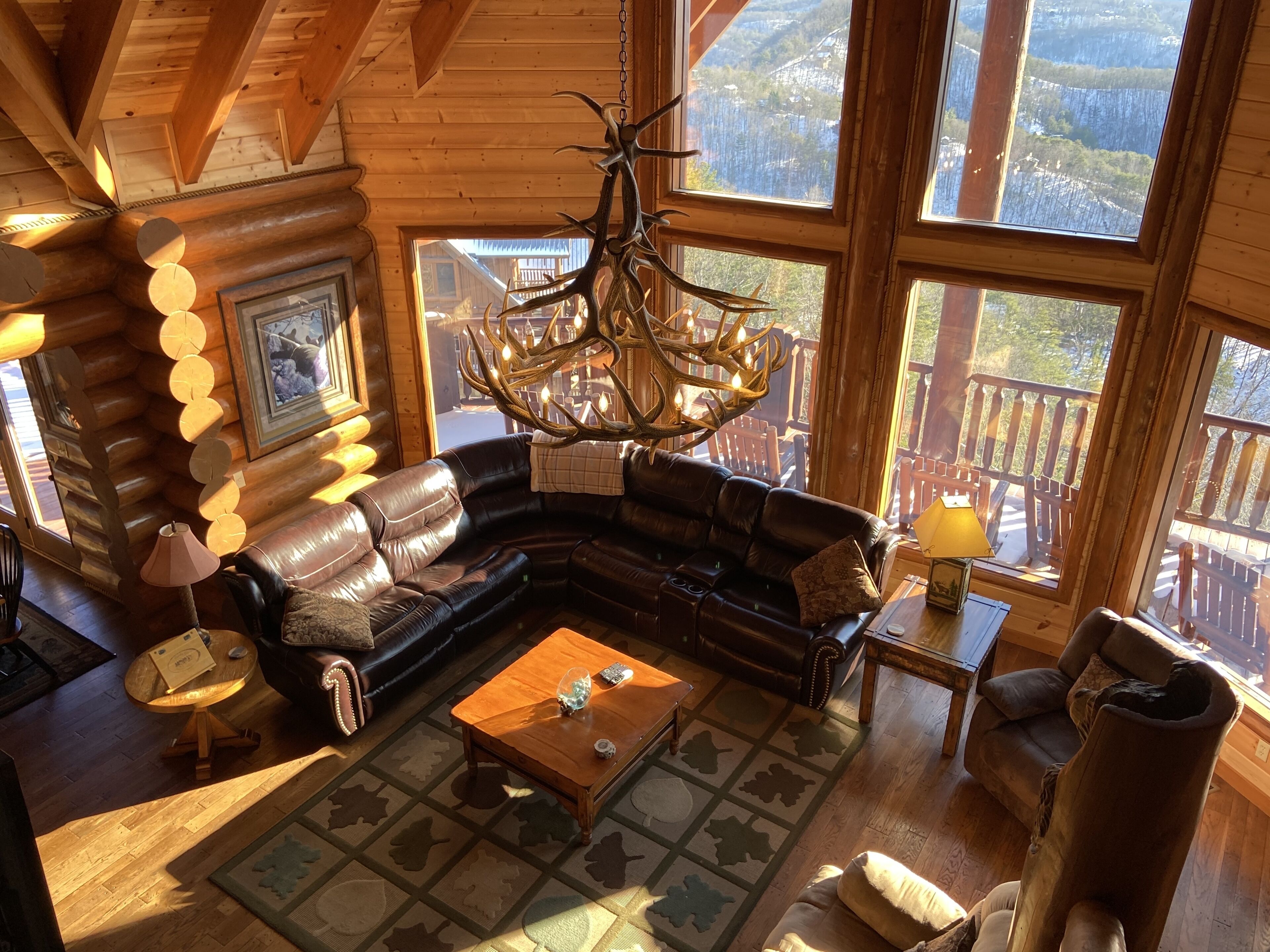 A cozy cabin interior with a leather sofa, wooden coffee table, and a chandelier. Large windows offer a view of the surrounding forest.