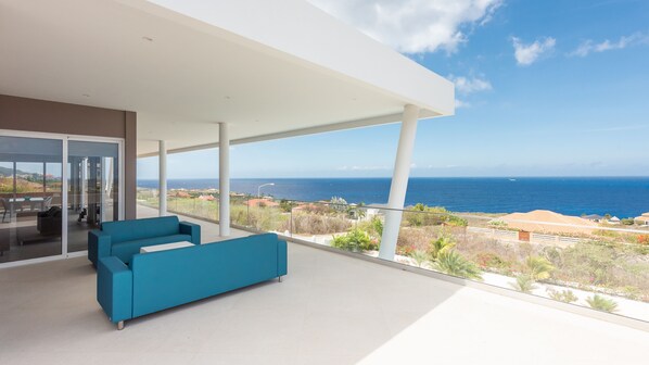 A modern balcony with a blue sofa, large glass doors, and a view of the sea and distant buildings.