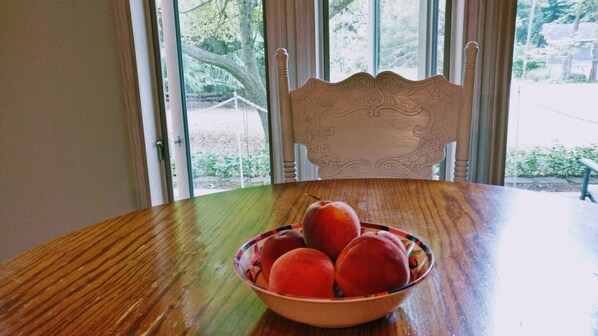 Una mesa de comedor de madera con un cuenco de fruta frente a una ventana que ofrece vistas a árboles y una valla.
