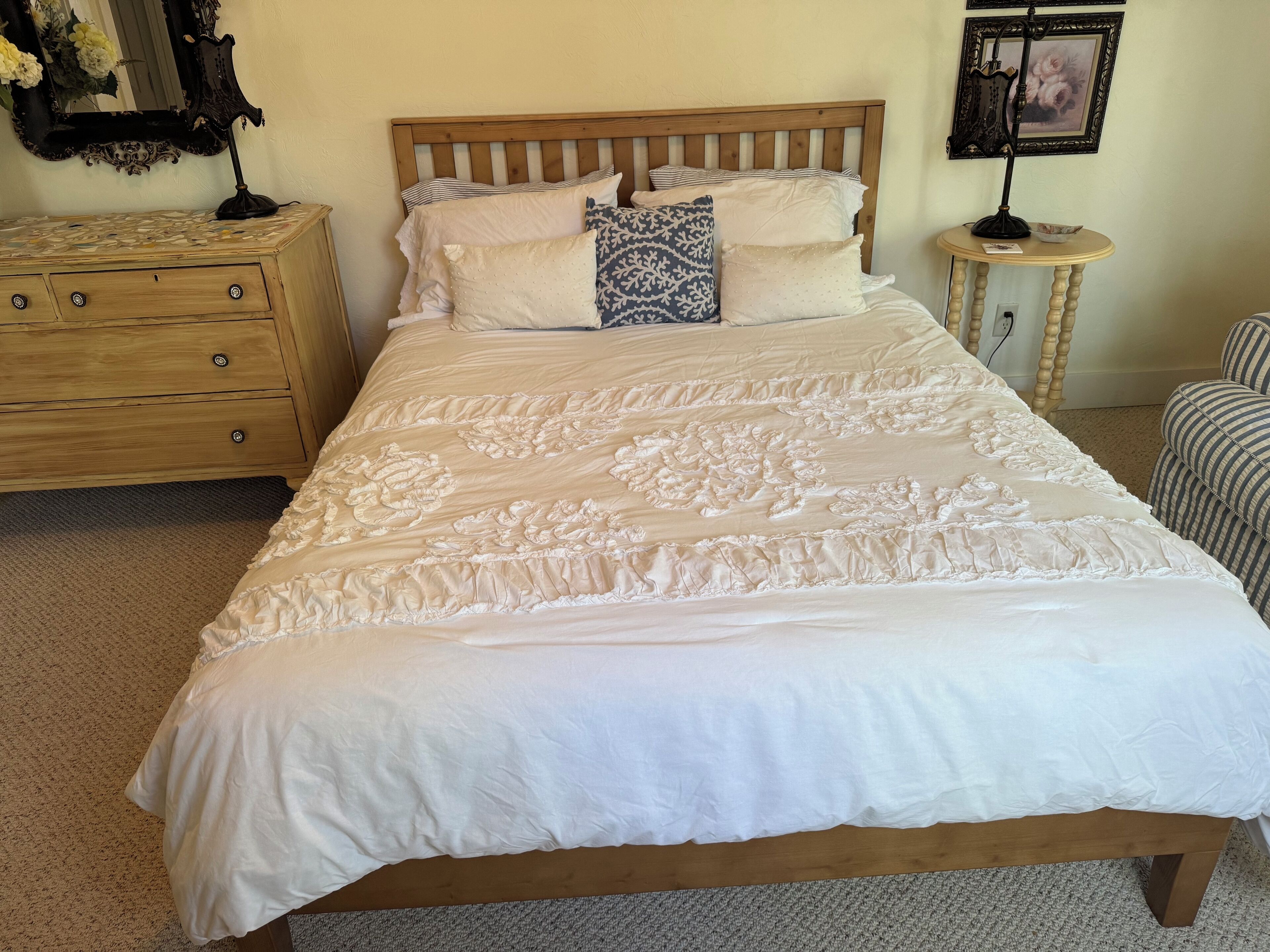 A neatly made bed with a wooden headboard, a wooden nightstand with drawers, a small round table with a lamp, and a framed picture on the wall.