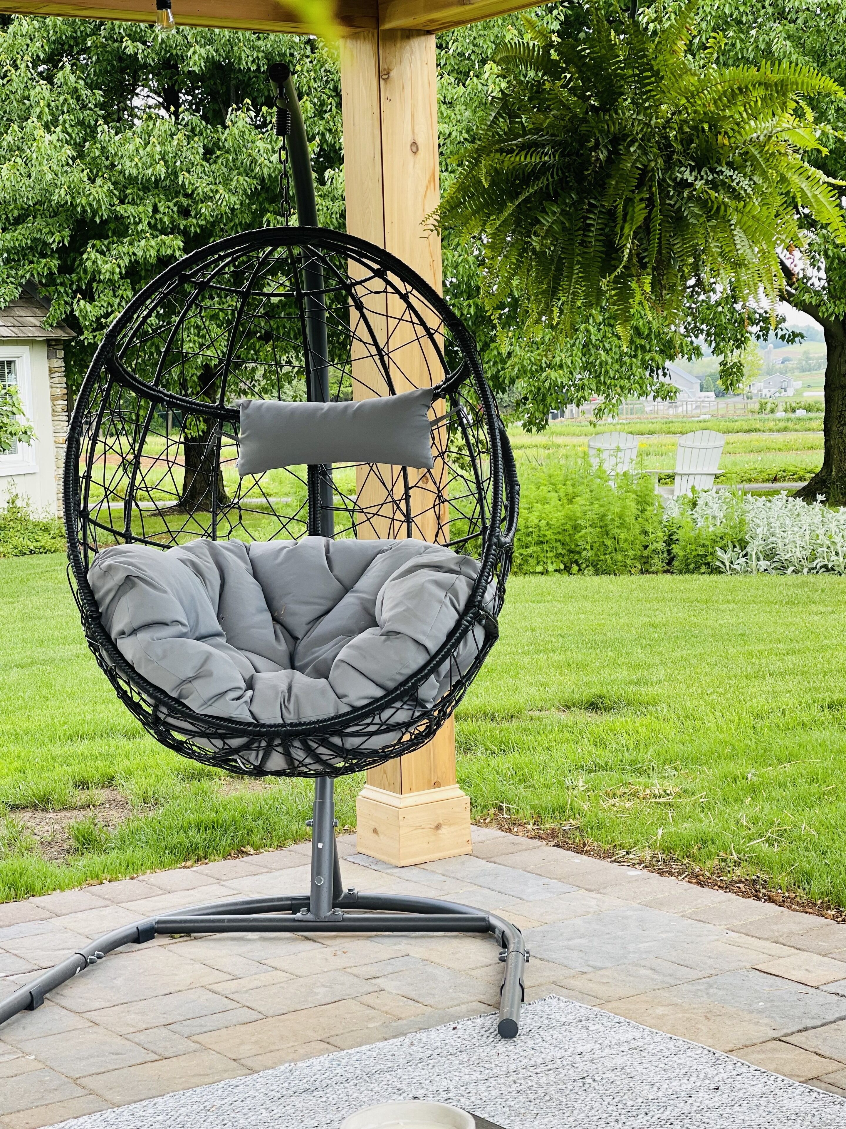 Una silla colgante con asiento y respaldo acolchados, soportada por una estructura de metal, ubicada en una zona pavimentada con césped y árboles al fondo.