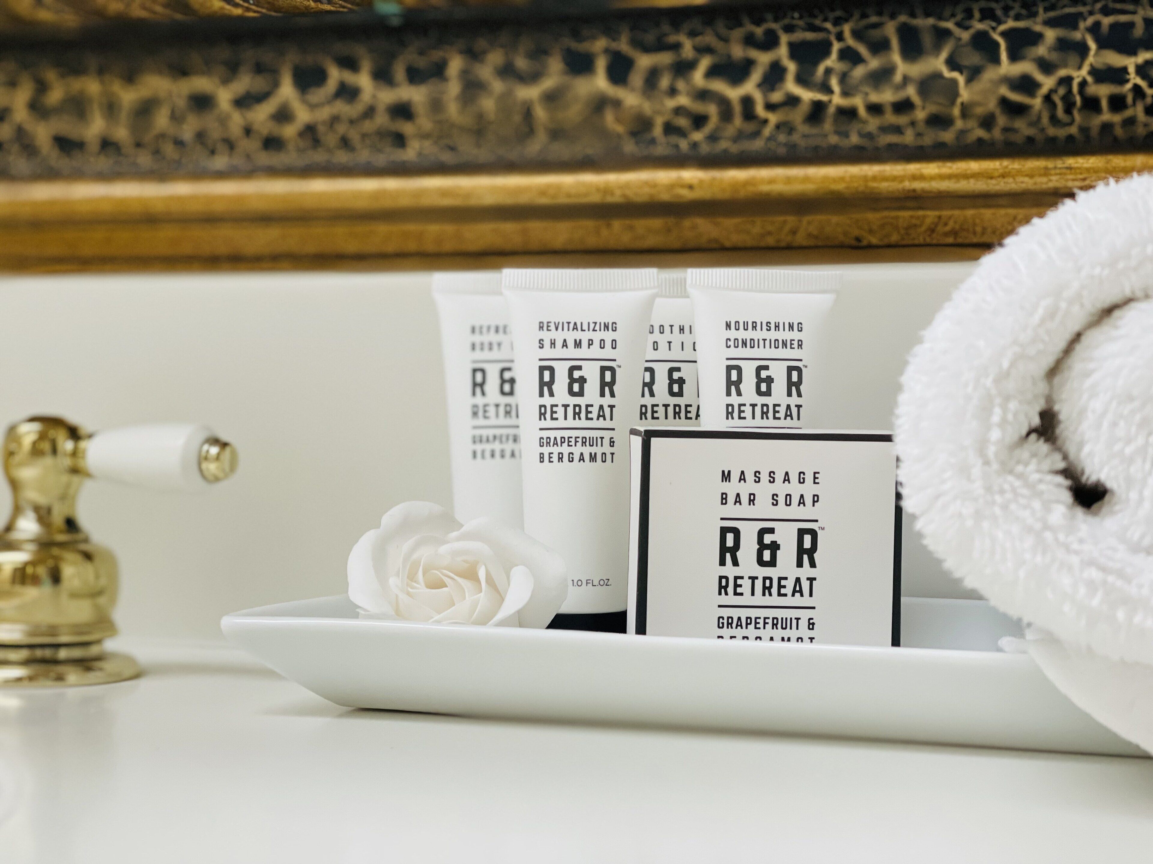 A tray with R&R Retreat products, including shampoo, conditioner, and bar soap, alongside a white rose and a gold faucet.