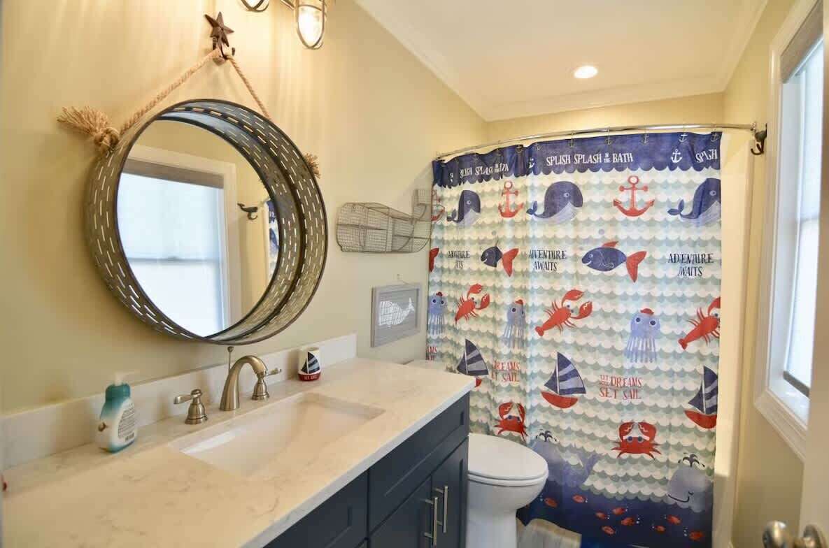 A bathroom with a nautical-themed shower curtain, a round mirror, and a sink with a towel dispenser.