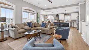 Ein geräumiges Wohnzimmer mit einem Ecksofa, einem Couchtisch und einem blauen Sessel. Es gibt eine Küchenzeile mit weißen Schränken sowie einen Essbereich mit Barhocker.