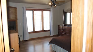 A bedroom with a large window, wooden floor, a bed, a dresser, and a ceiling fan.