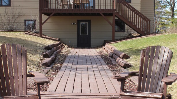 A wooden deck leading to a house with a front porch and two Adirondack chairs.