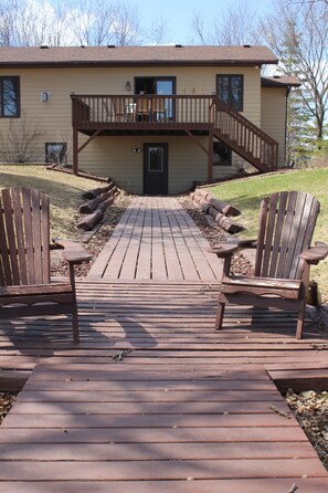 A wooden deck leading to a house with a front porch and two Adirondack chairs.