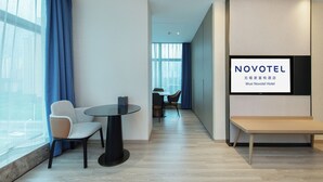 Moderni hotellihuone, jossa on suuri ikkuna, oleskelualue pöydällä ja tuoleilla sekä seinälle asennettu televisio, jossa näkyy hotellin nimi.