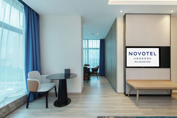 A modern hotel room with a large window, a seating area with a table and chairs, and a wall-mounted TV displaying the hotel name. - Novotel Wuxi (Wuxi)