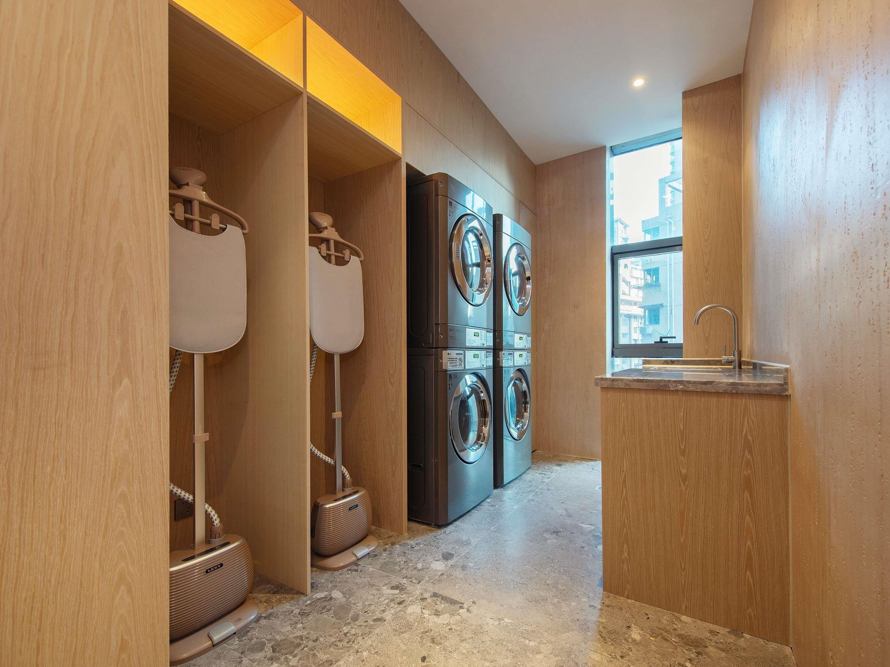 Una sala de bugaderia amb piles de rentadores i assecadores, un rentapousa i una finestra amb vista a edificis.