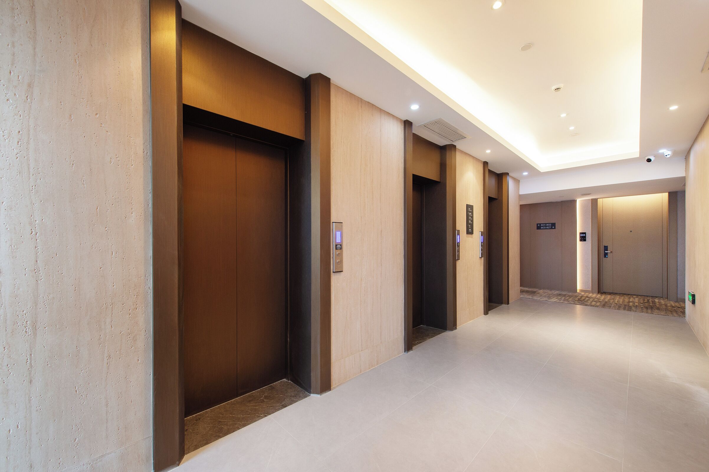 A modern hotel corridor with elevators, a clean floor, and a ceiling with recessed lighting.
