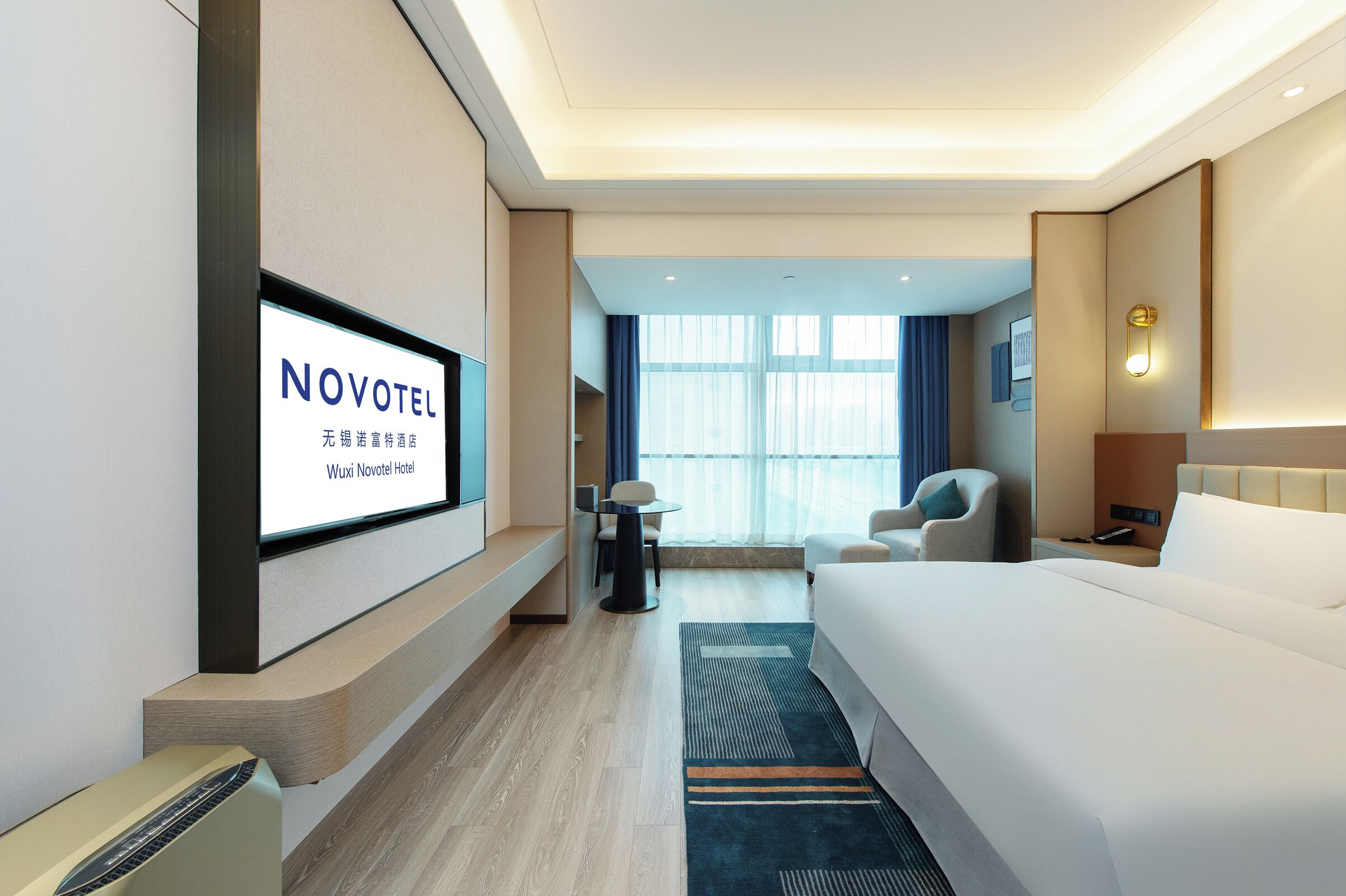 Ein Hotelzimmer mit Bett, Wand-Fernseher, Schreibtisch, Stuhl und einem fensterbepflanzten Fenster.