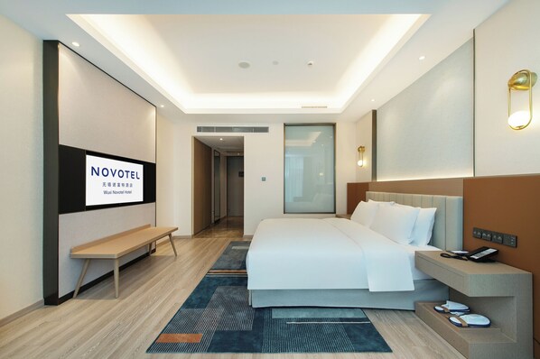 A modern hotel room with a bed, bedside table, and a TV displaying 'NOVOTEL'. - Novotel Wuxi (Wuxi)