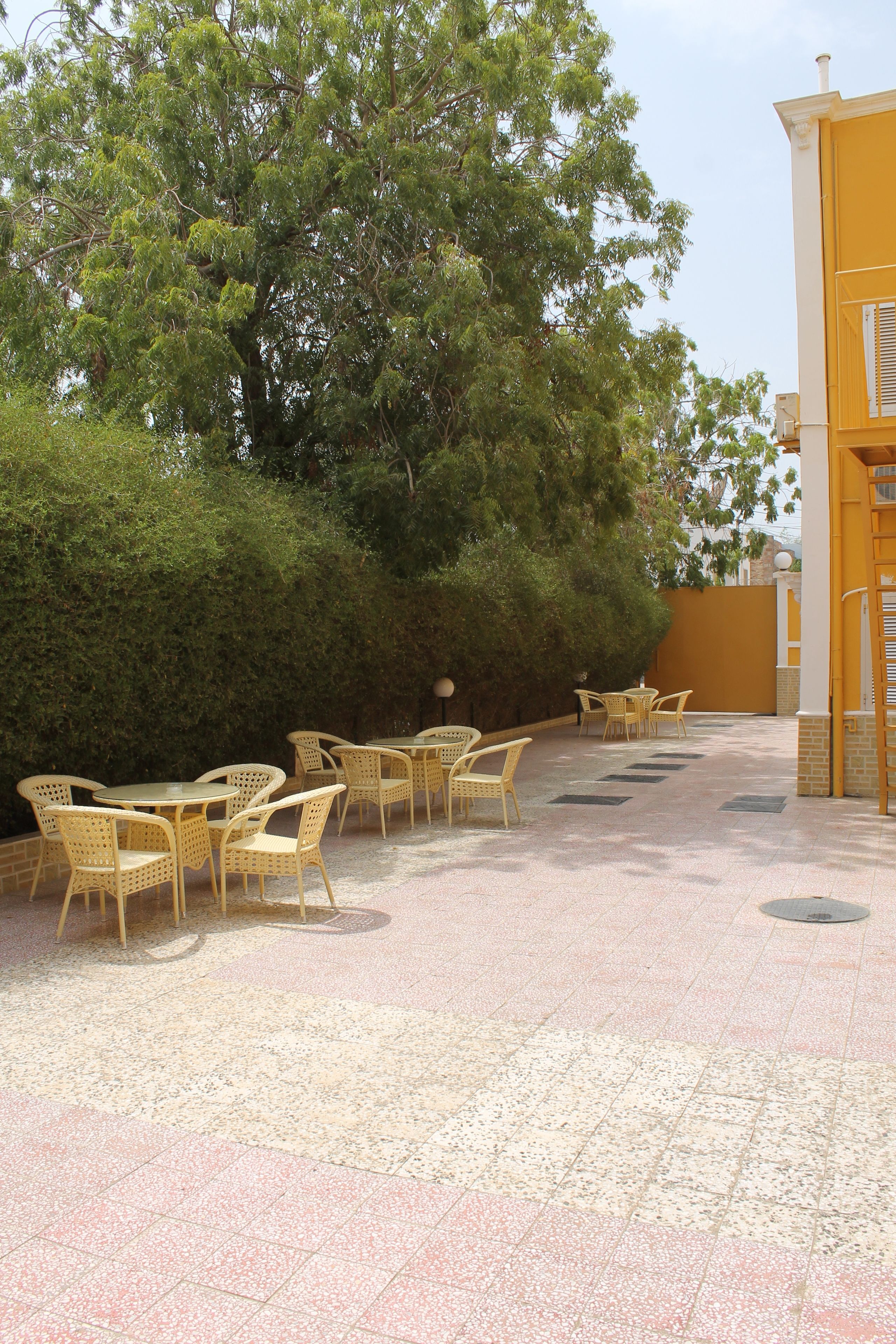 Un patio al aire libre con mesas y sillas de mimbre, rodeado de follaje y un edificio amarillo.