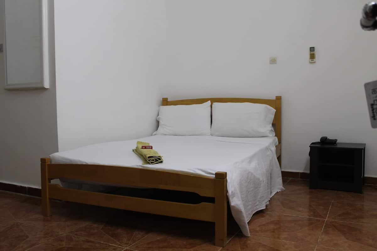 Una cama individual con estructura de madera y ropa de cama blanca, una mesita de noche pequeña con un teléfono, y un aplique de pared.