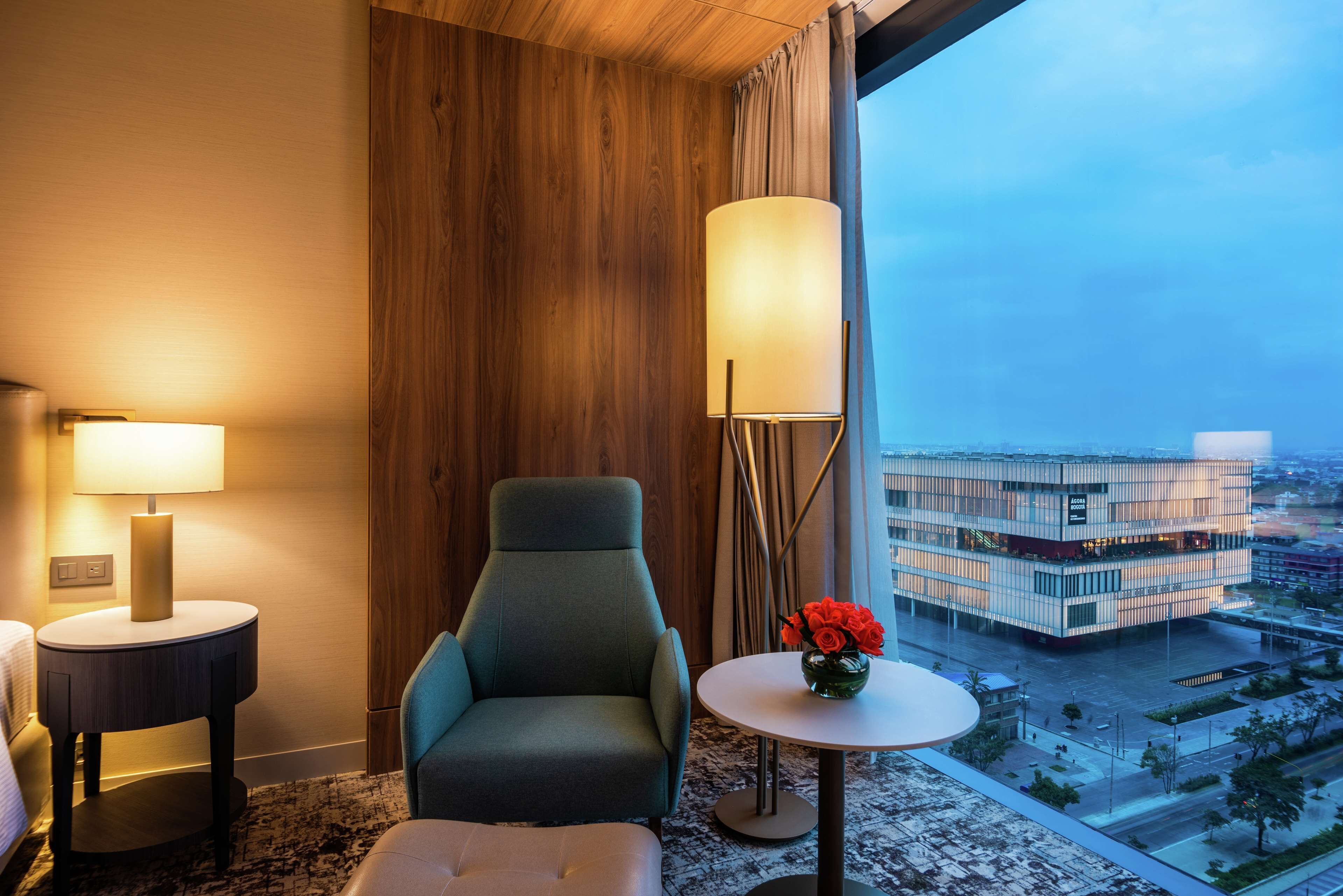 A hotel room with a green armchair, a round table with a vase of red flowers, a standing lamp, and a view of a modern building through large windows.