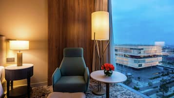 A hotel room with a green armchair, a round table with a vase of red flowers, a standing lamp, and a view of a modern building through large windows.