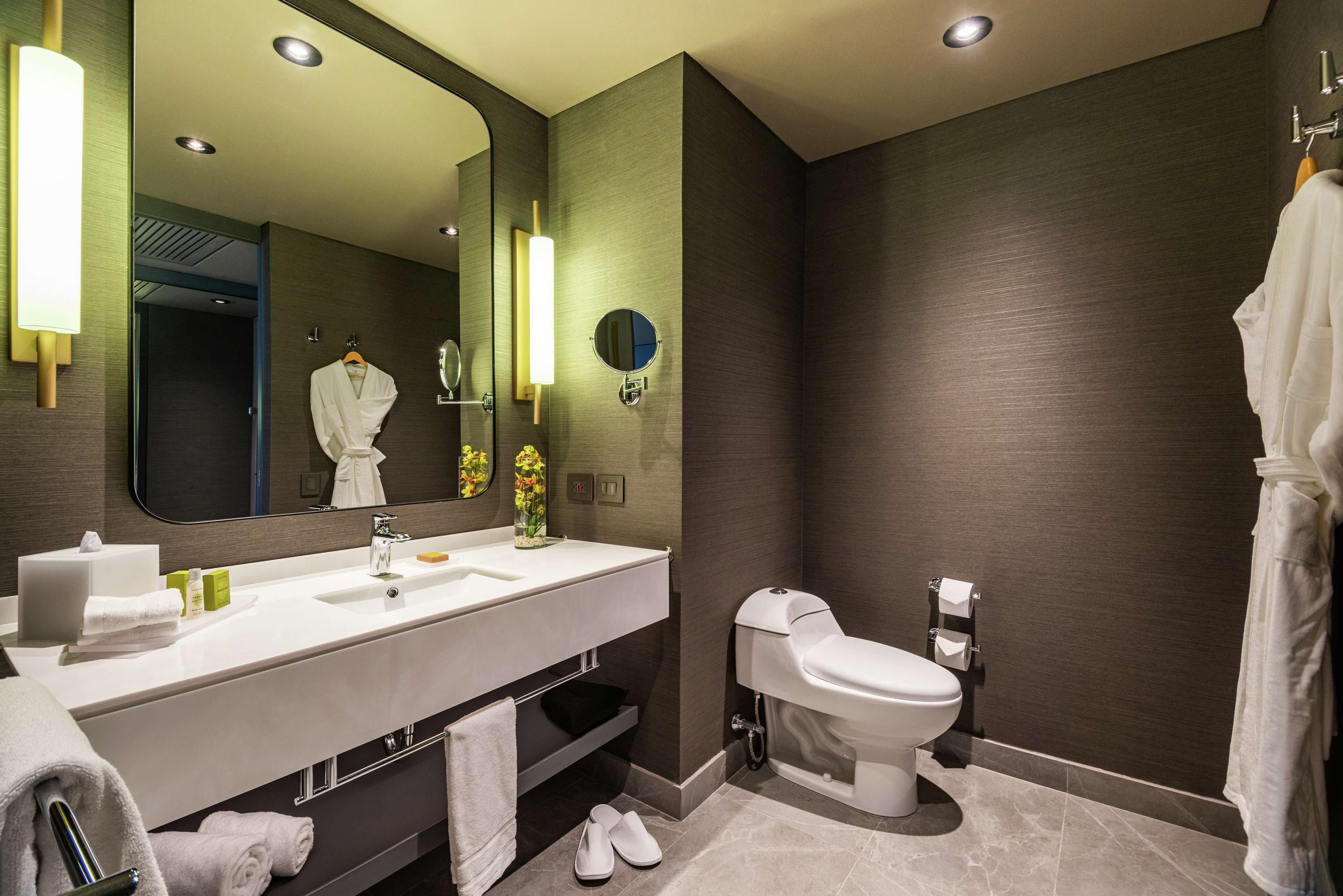 A modern bathroom with a large mirror, white sink, and toilet. There are towels and toiletries on the counter.