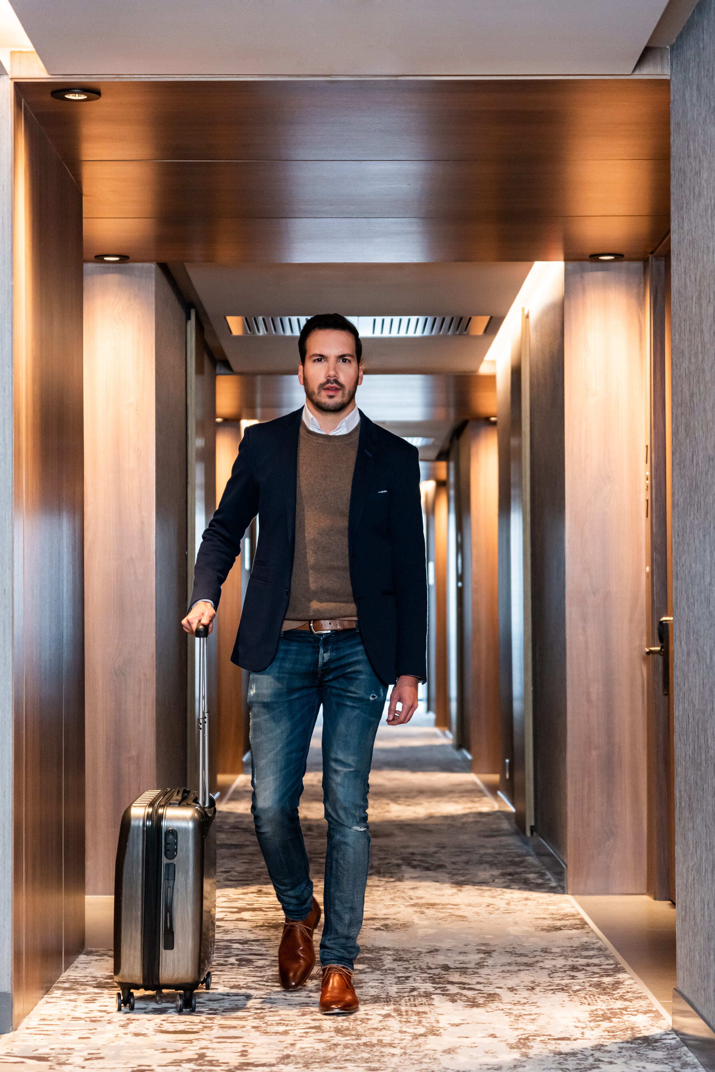 Um homem em um corredor de hotel com paredes de madeira e piso coberto de carpete, carregando uma mala.