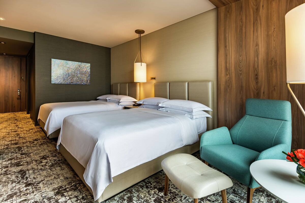 Habitación de hotel con dos camas, un sillón verde azulado, un banco blanco y un cuadro en la pared.
