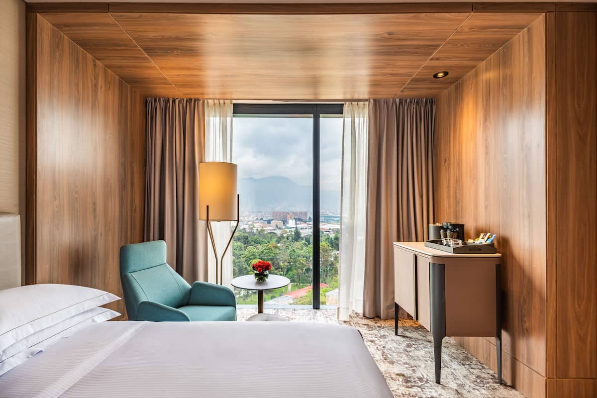 Habitación de hotel con cama, una silla, una mesita de noche con lámpara y vistas a la ciudad a través de amplias ventanas.