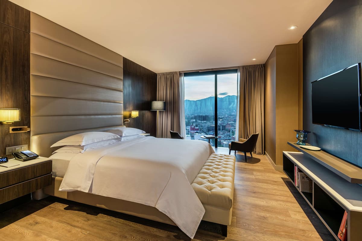 Una habitación de hotel moderna con una cama grande, un televisor, un escritorio con teléfono y vistas a un paisaje urbano a través de un ventanal.