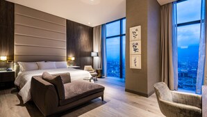 Une chambre d’hôtel moderne avec un grand lit, un canapé, un fauteuil et une vue sur la ville par les fenêtres.