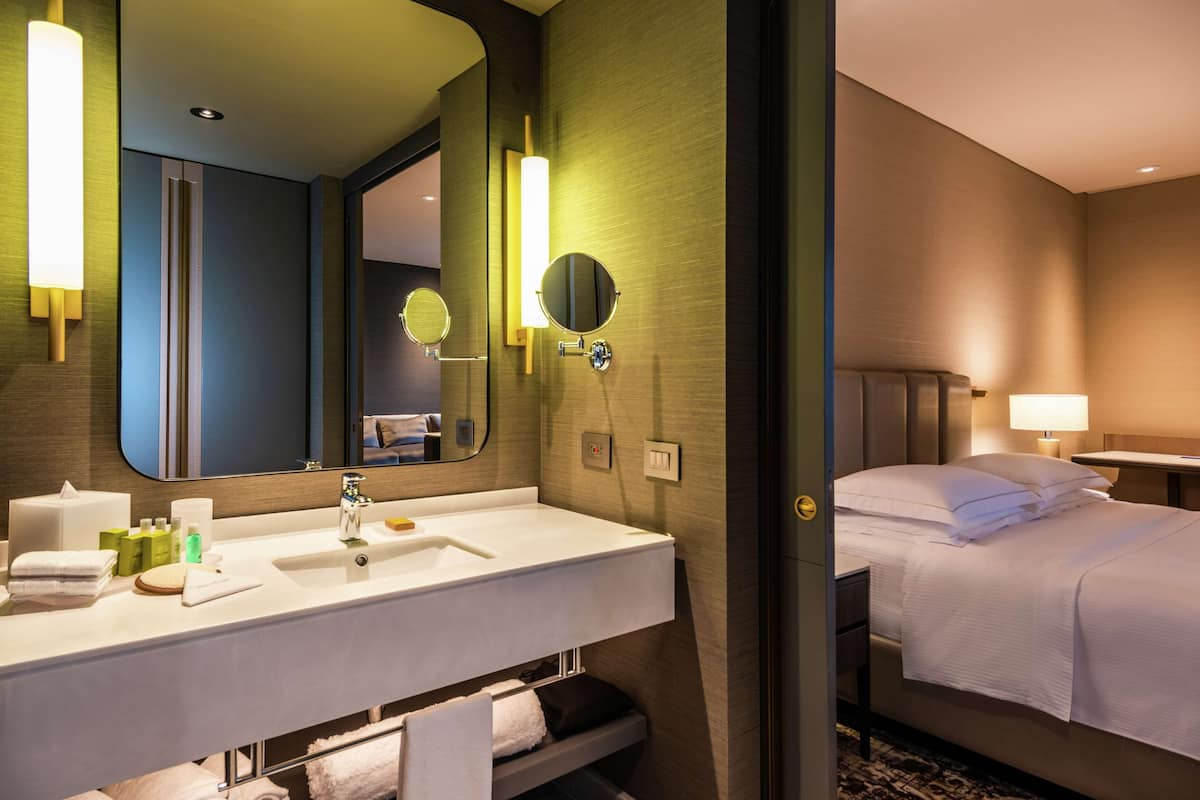 Habitación de hotel con un espejo grande, un lavamanos blanco, una cama con ropa de cama blanca y una mesita de noche con lámpara.
