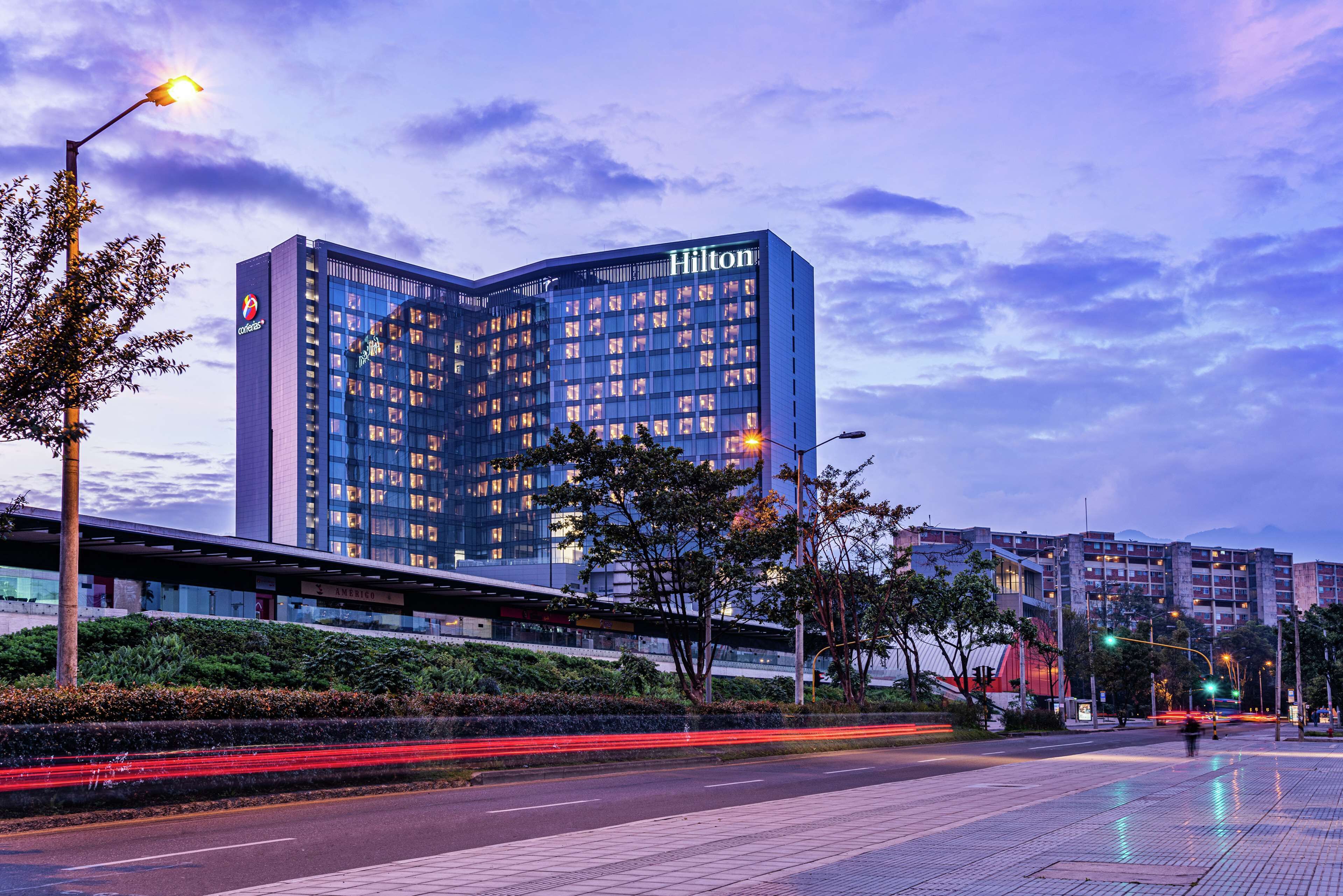 Un hotel moderno con un letrero llamativo de Hilton, rodeado de follaje y una calle urbana bien iluminada.