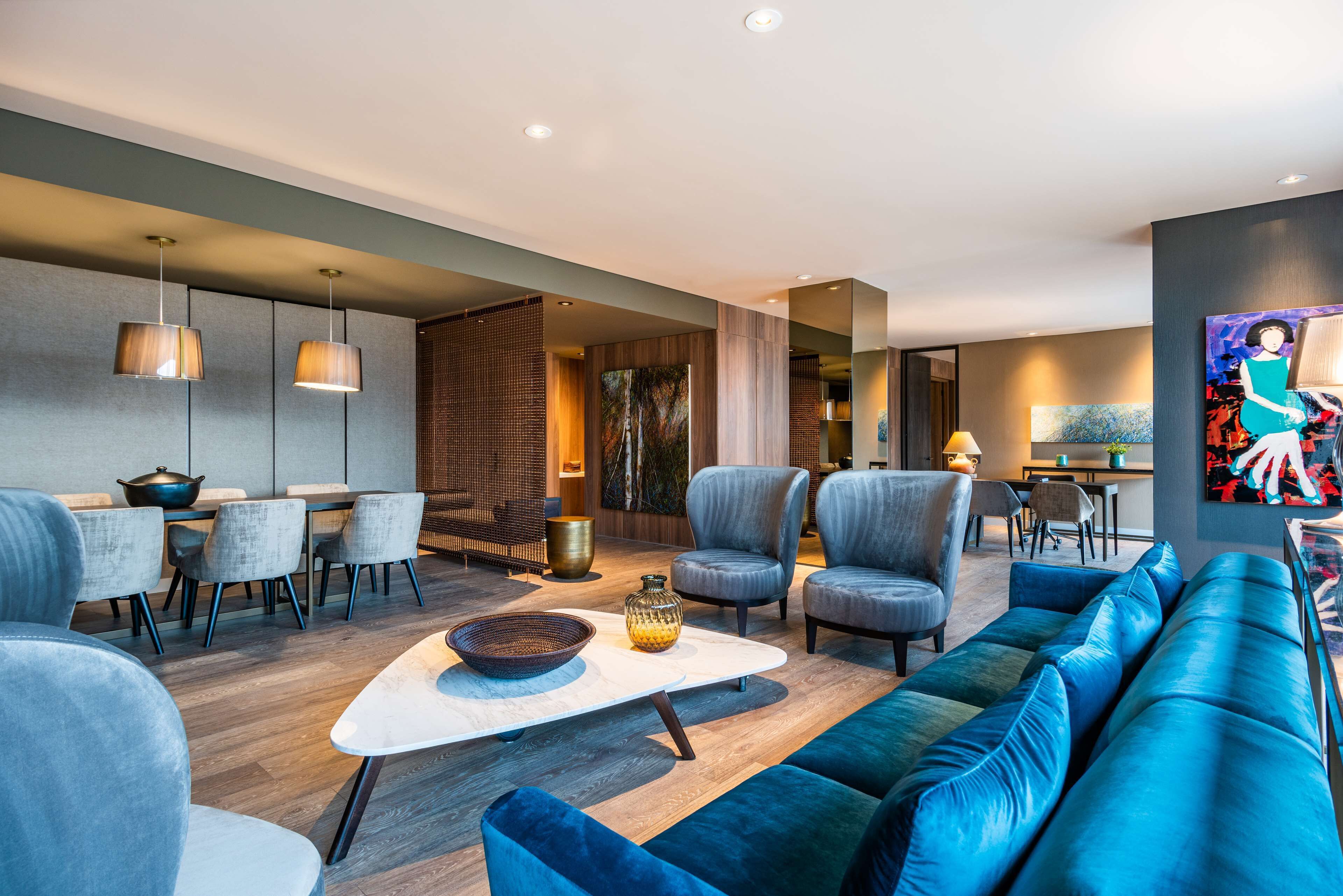 A modern living room with a blue sofa, grey armchairs, a white coffee table, and a dining area with a table and chairs.