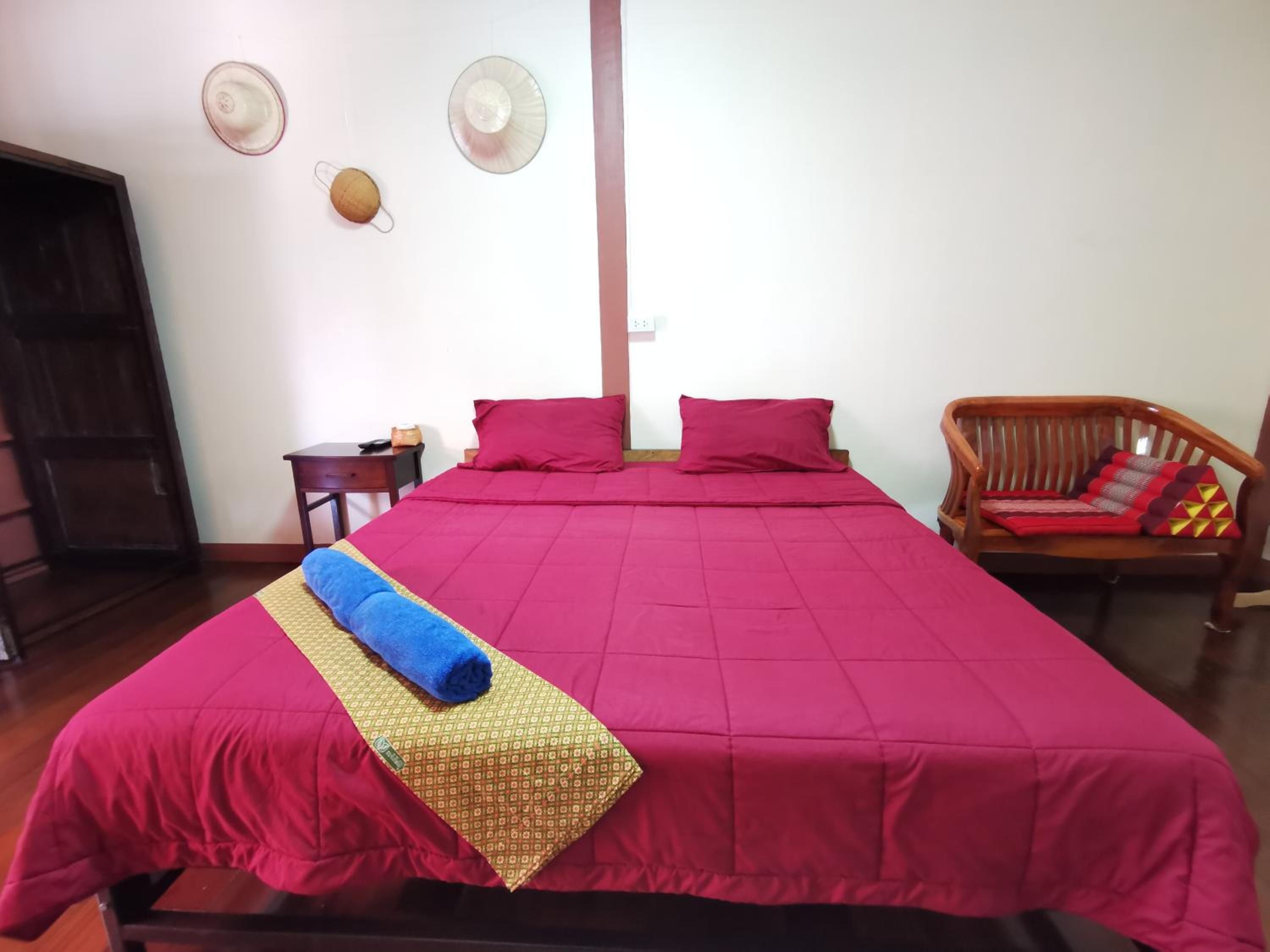A room with a large bed, a wooden chair, a small table, and a wall-mounted fan.