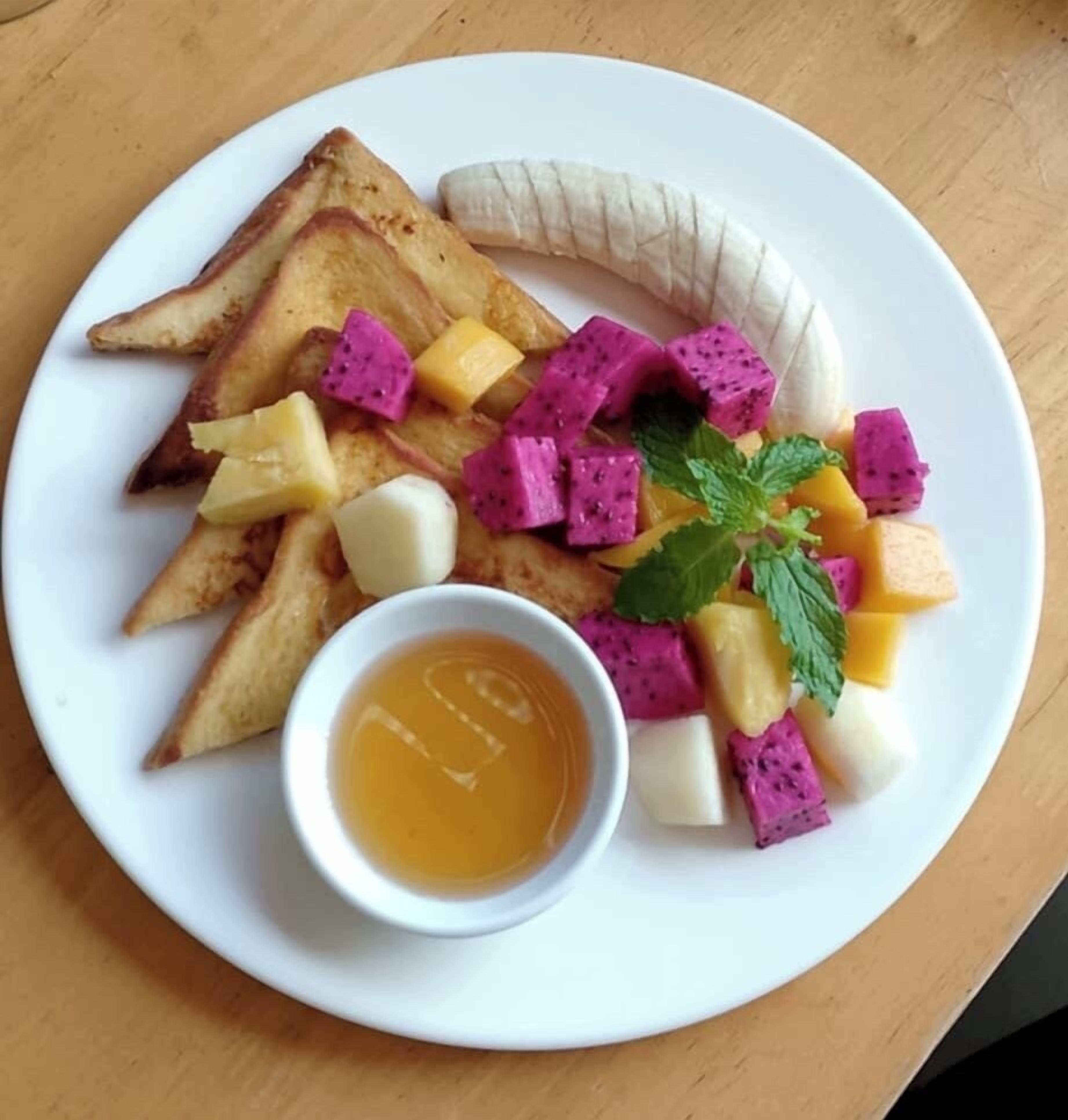 Een bord met toast, banaan, pitahaya, mango en een klein schaaltje siroop.