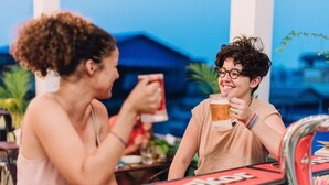Dos personas sentadas en una mesa, cada una con una bebida en la mano, con un letrero rojo que dice "Anger" y un cielo azul de fondo.