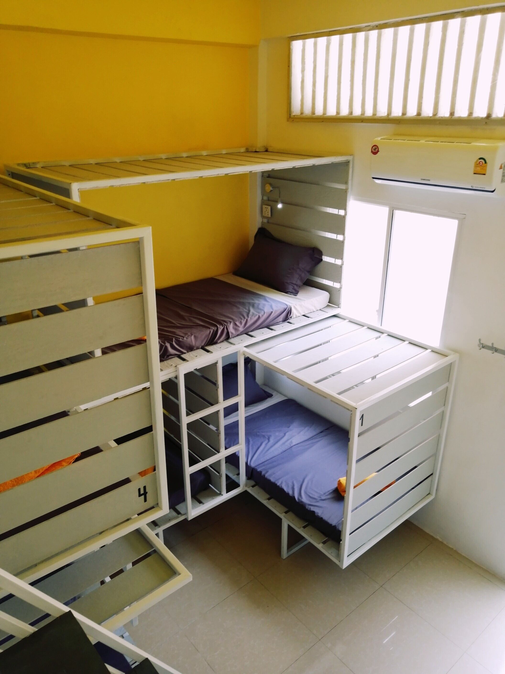 A dormitory room with bunk beds, a window with blinds, and a wall-mounted air conditioner.