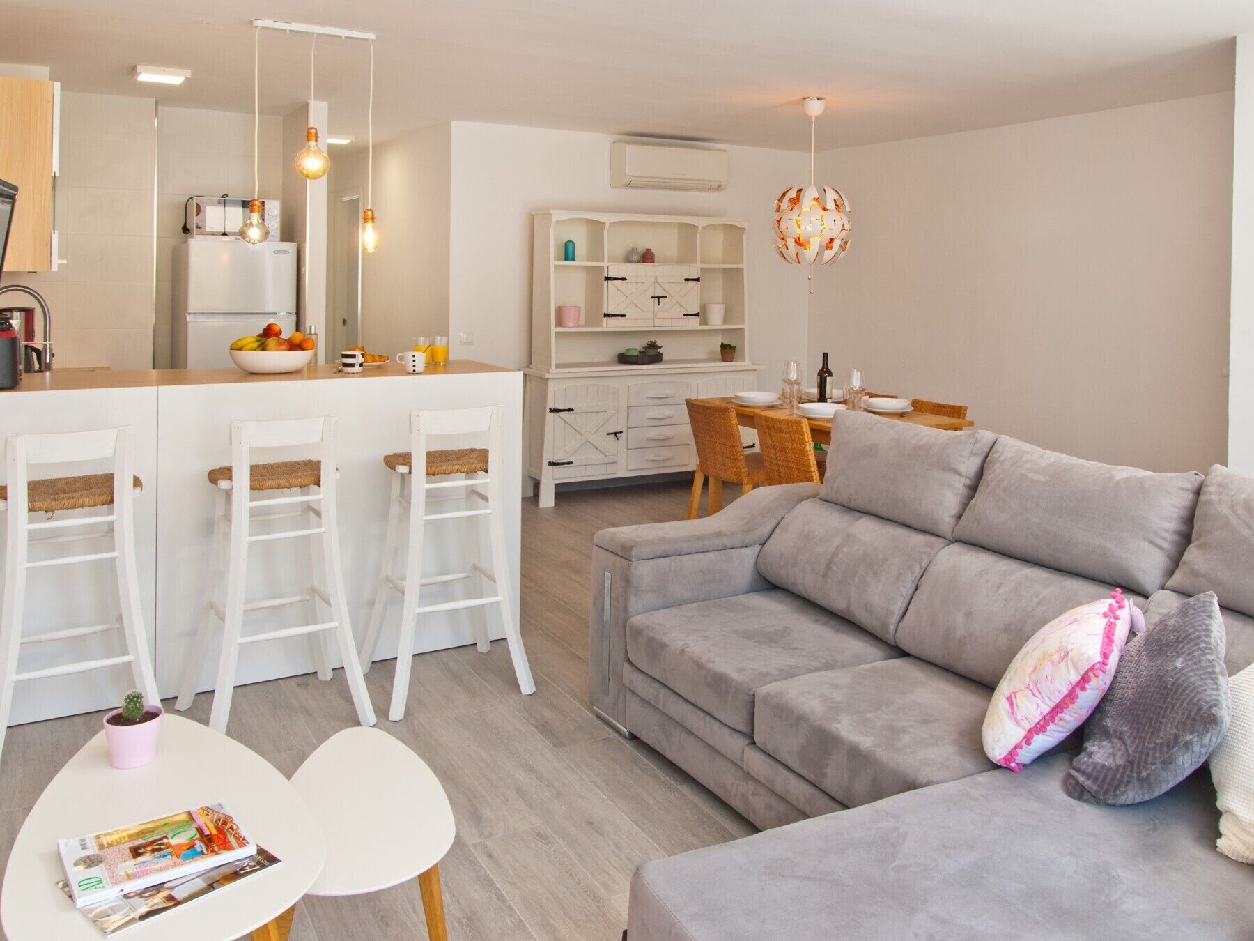 Een moderne woonkamer met een grijze bank, een klein wit tafeltje en een keuken met een koelkast, barstoelen en een eettafel.