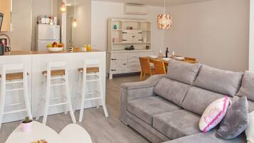 Een moderne woonkamer met een grijze bank, een klein wit tafeltje en een keuken met een koelkast, barstoelen en een eettafel.