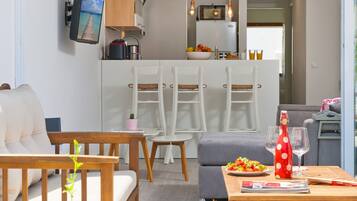 Een moderne woonkamer met een bank, een houten tafel en een eethoek met stoelen. Achtergrond is een keuken met een koelkast en een magnetron.