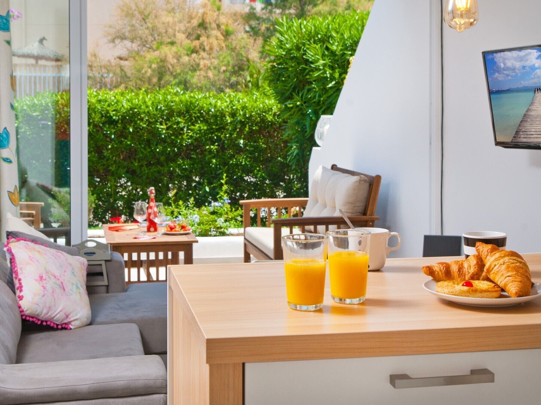 Een moderne kamer met een houten eettafel, twee glaasjes sinaasappelsap, een bord met croissants en een klein gebakje, en uitzicht op een weelderige tuin door een groot raam.