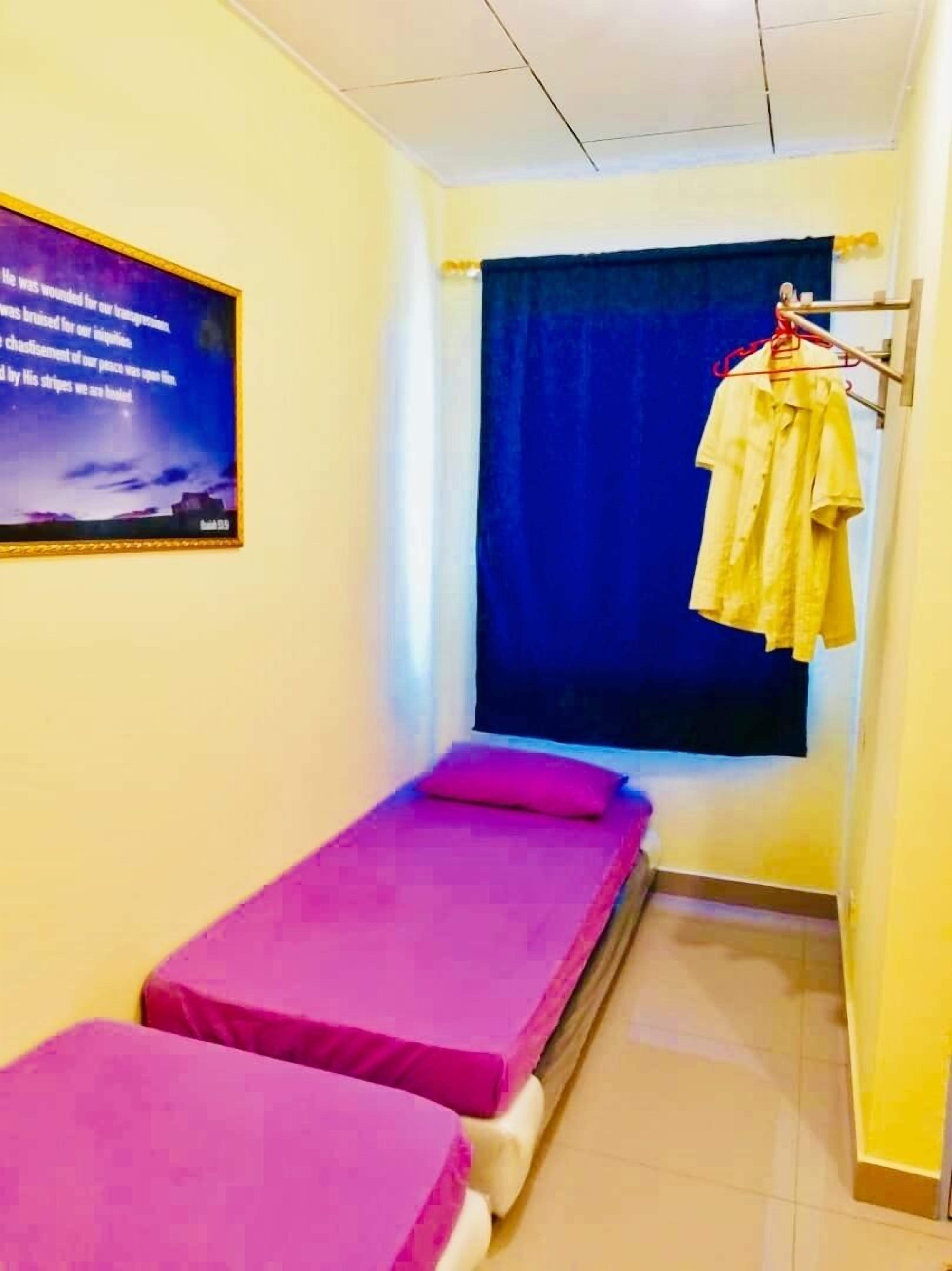 A small, narrow room with two single beds, a window with a blue curtain, and a wall-mounted picture.