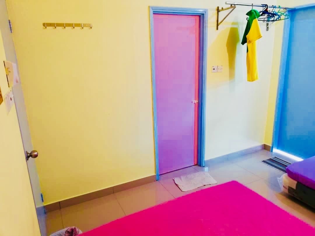 Une pièce avec un tapis rose, une porte dotée d’un cadre bleu et un étendoir fixé au mur, sur lequel sont suspendus des vêtements.