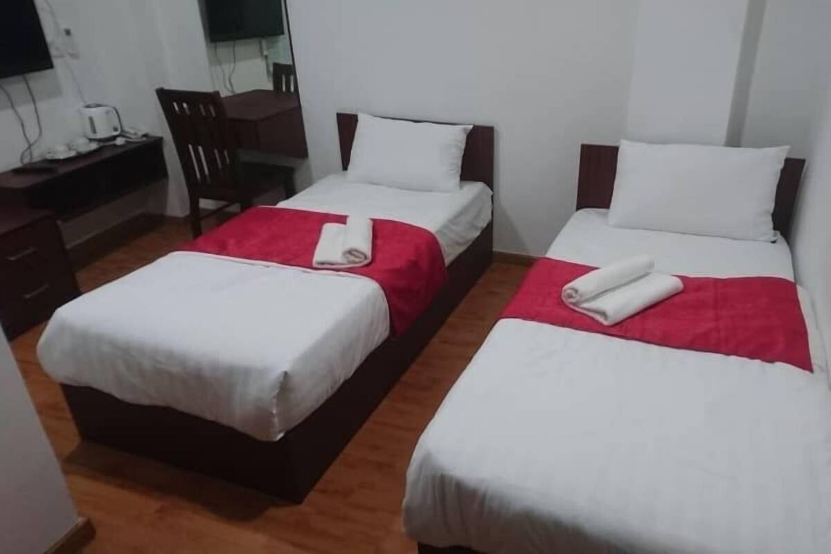 Habitación de hotel con dos camas individuales, cada una con ropa de cama blanca y detalles rojos, un escritorio de madera con una silla y un televisor de pantalla plana montado en la pared.