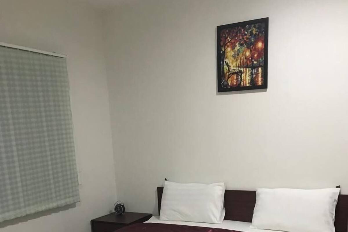 Una cama bien hecha con sábanas blancas y una toalla doblada, un cabecero oscuro y una mesita de noche con una lámpara.