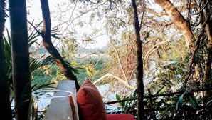 A glass of water on a table with a red cushion in a shaded outdoor area with trees and a view of a natural landscape.