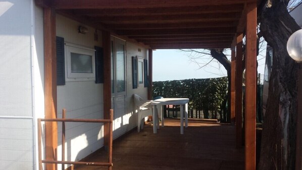 Un espace extérieur abrité, doté d’un plancher en bois, d’une table blanche et d’un banc.