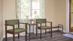 Una habitación con dos sillas de madera con cojines verdes, una mesita de noche con una lámpara y una ventana con cortinas estampadas.