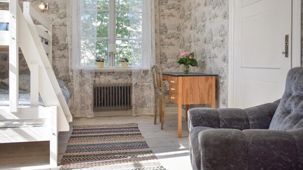 Una habitación con una cama litera, un escritorio de madera, una silla, un sofá gris, un tapete estampado y una ventana con plantas en macetas.