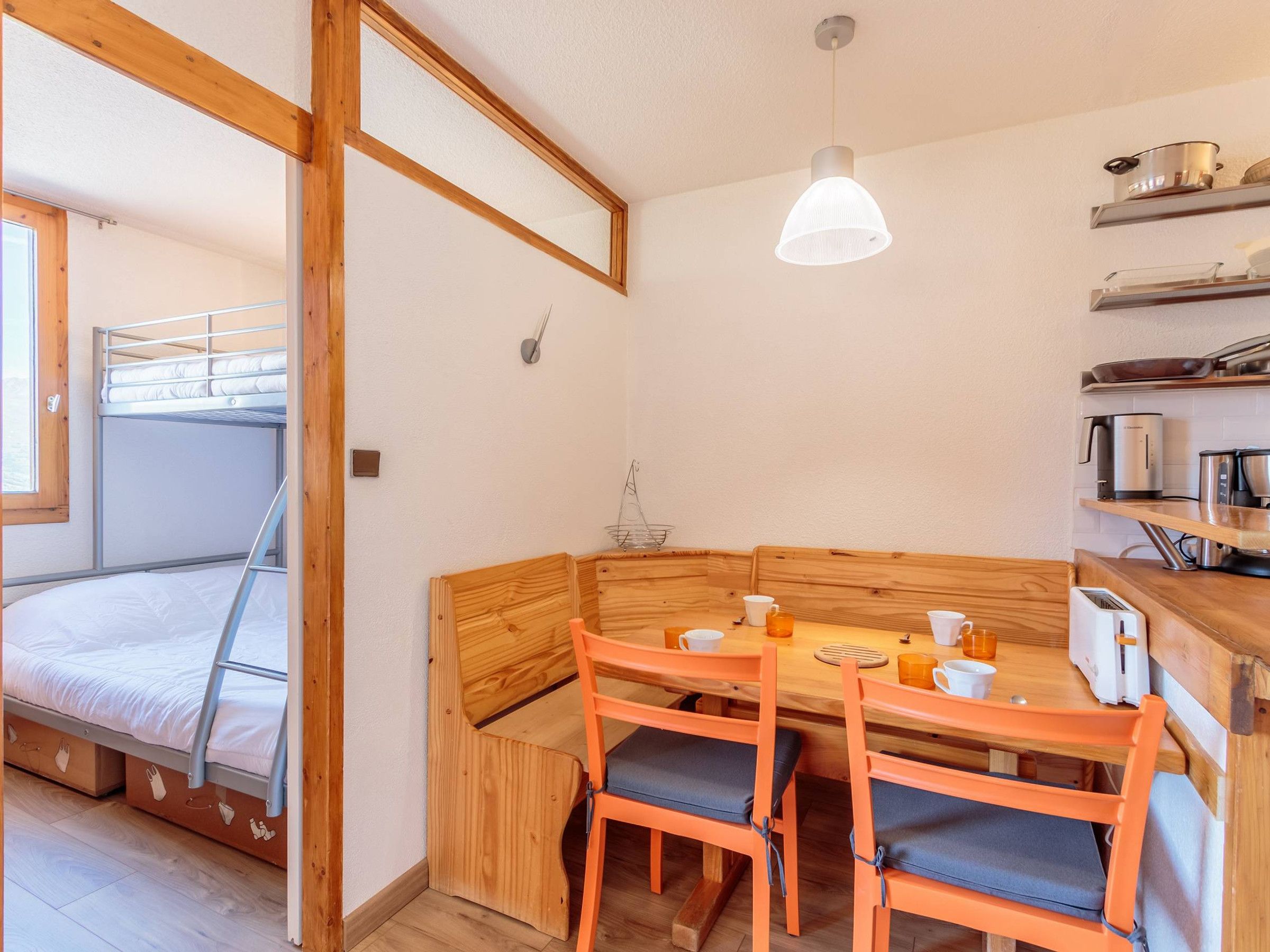 A compact room with a bunk bed, wooden dining table with chairs, a hanging light fixture, and kitchen appliances on shelves.