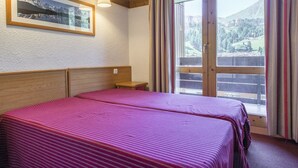 A hotel room with two beds, a wooden headboard, a small bench, a framed picture on the wall, and a large window with curtains. - 43m² Apartment in Plagne 1800, pets allowed, parking included (La Plagne 1800)