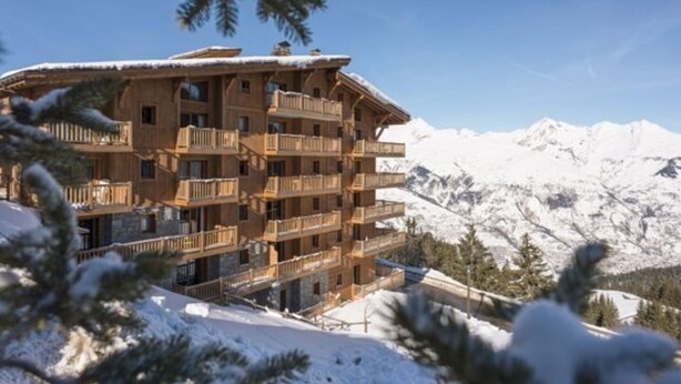 A multi-story wooden building with balconies, surrounded by snow-covered mountains and pine trees.