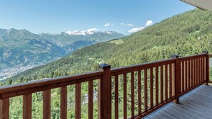 A wooden balcony with a view of a mountainous landscape, including snow-capped peaks and dense forest.